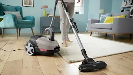 Someone is vacuuming the light floor in a living room with a cylinder vacuum cleaner; a yellow sofa and white table are in the background.