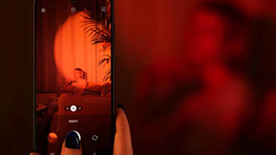 Een smartphone toont een foto van een persoon op een bank met een lamp en planten op de achtergrond, tegen een rode achtergrond.