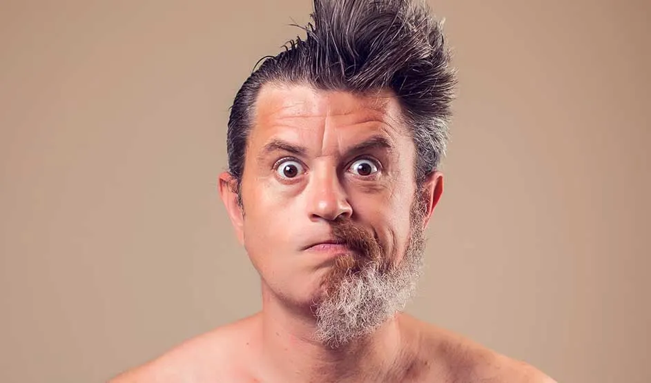 A man stares in surprise at the camera; his hairstyle and beard are half neatly groomed and half wild and messy.