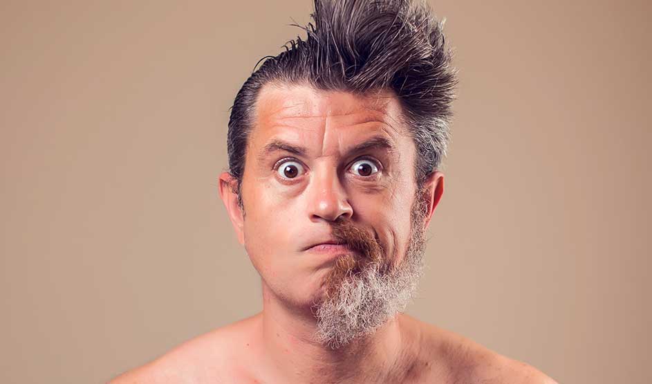 Een man kijkt verbaasd in de camera; zijn kapsel en baard zijn half strak verzorgd en half wild en ongekamd.