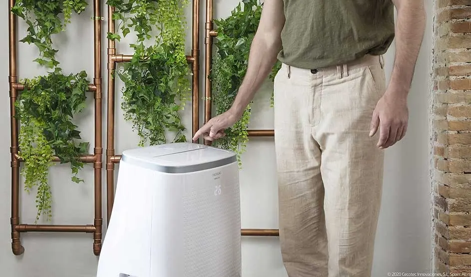 A man in light trousers is using a white Cecotec air conditioner, which stands near green plants and copper pipes in a stylish, modern room.