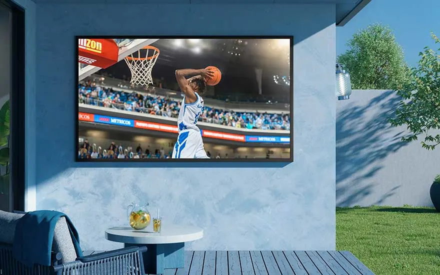 Een buitenruimte met een grote tv aan een blauwe muur, waarop live basketball wordt uitgezonden, naast een modern terrasmeubel.