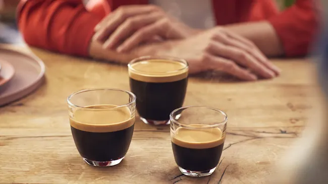 Drie espresso's in glazen bekers op een houten tafel, met een persoon in een rood vest op de achtergrond.