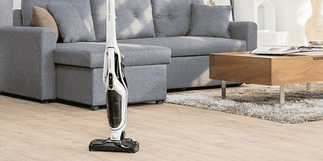 Een witte stofzuiger staat op een houten vloer in een woonkamer met een grijze bank en een houten tafel op een grijs tapijt.