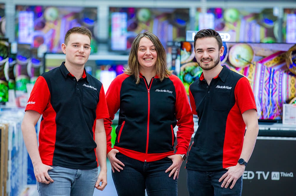Three employees in black and red uniforms stand smiling in front of display televisions in an electronics store.