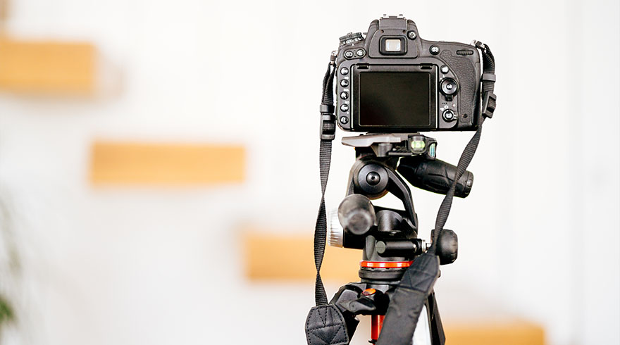 Een zwarte camera staat op een statief, met een witte muur en houten planken op de achtergrond.