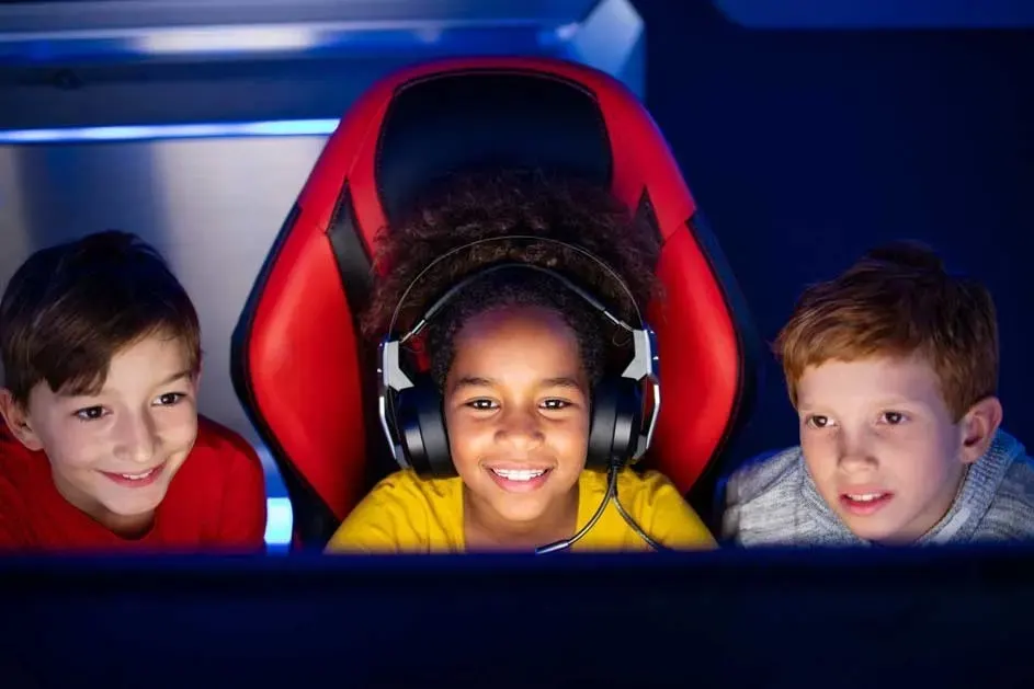 Three children sit together behind a screen. The middle child wears a large gaming headset and sits in a red game chair. No brand visible.