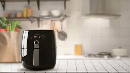 Een zwarte airfryer op een wit aanrecht met een papieren zak groenten ernaast, in een keuken met planken en gereedschap.