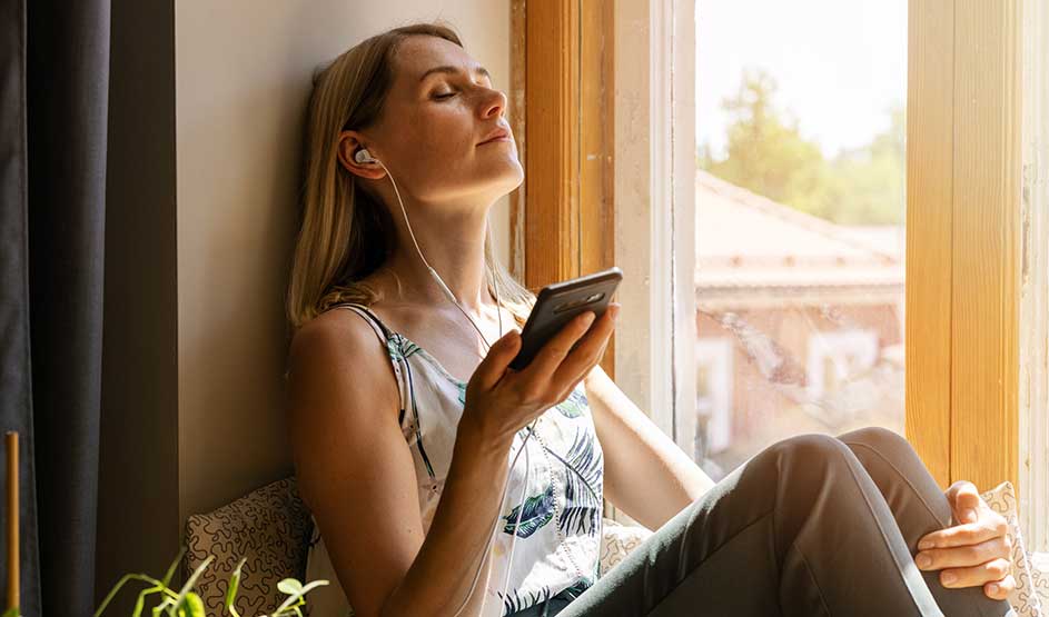 Vrouw leunt ontspannen bij het zonnige raam, ogen gesloten, met bedrade oordopjes in en smartphone in de hand, genietend van muziek.