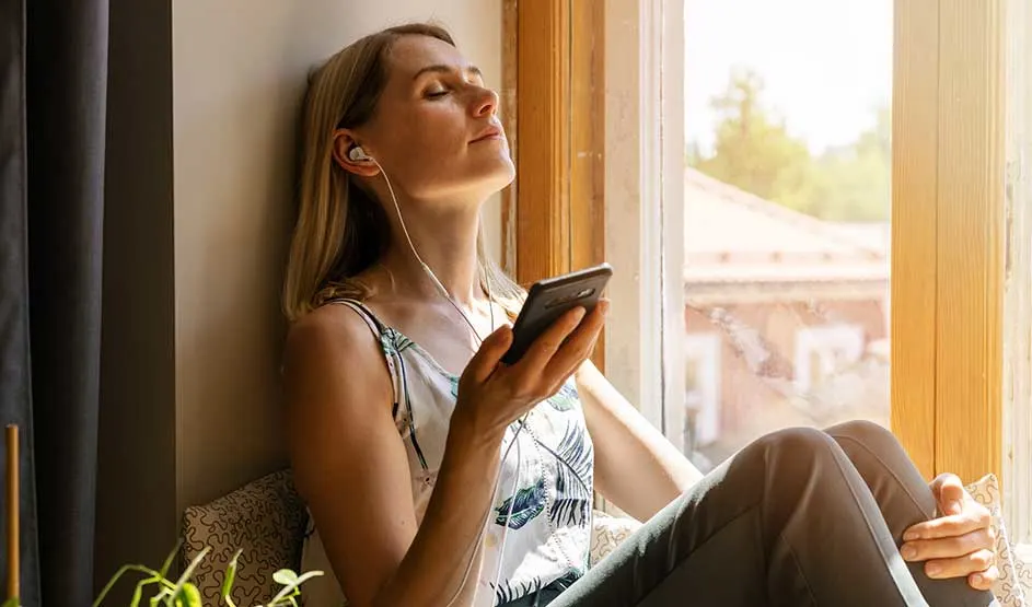 Vrouw leunt ontspannen bij het zonnige raam, ogen gesloten, met bedrade oordopjes in en smartphone in de hand, genietend van muziek.