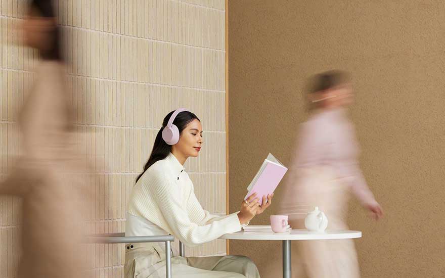 Woman with pink headphones reads calmly at a small white table as blurred passers-by move past her in soft tones.
