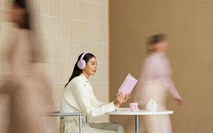 Woman with pink headphones reads calmly at a small white table as blurred passers-by move past her in soft tones.