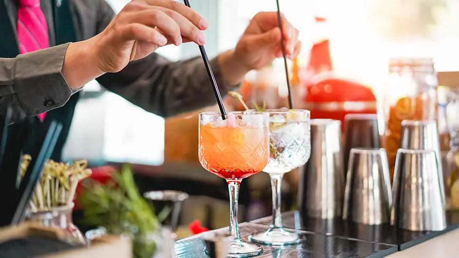 Een barman roert in twee cocktails met rietjes, naast cocktailshakers en garnering op een bar.