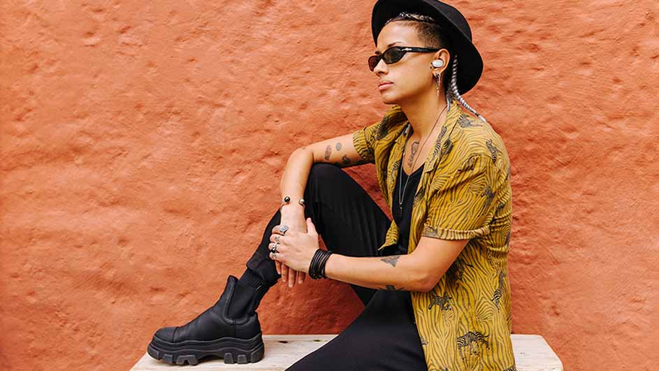 Een persoon met een hoed en zonnebril zit voor een oranje muur. Ze dragen een geel shirt met zebraprint en zwarte kleding.