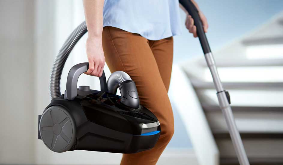 A person carries a compact black cylinder vacuum by its handle, hose and wand at their side while walking past stairs indoors.
