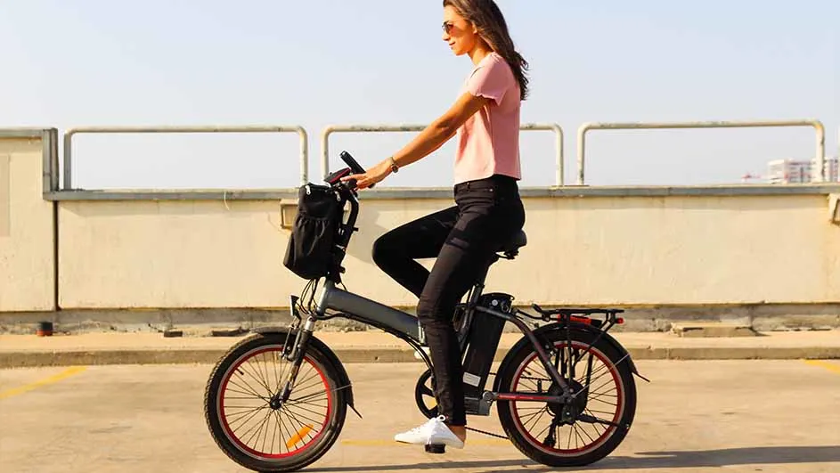 Vrouw op grijze elektrische fiets met zwarte tas. Ze draagt een roze shirt, zwarte broek en witte schoenen. Achtergrond: betonnen muur.