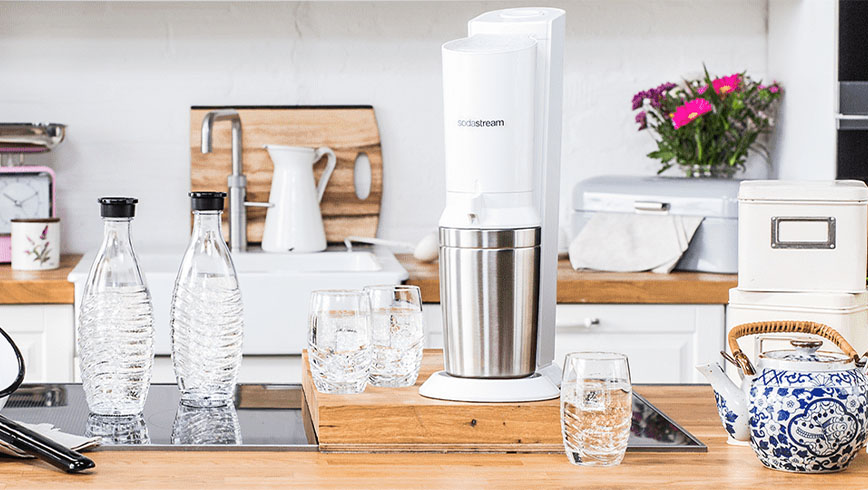 Keukenopstelling met Sodastream, flessen en glazen gevuld met water. Een blauw-witte theepot staat rechts, bloemen achteraan.