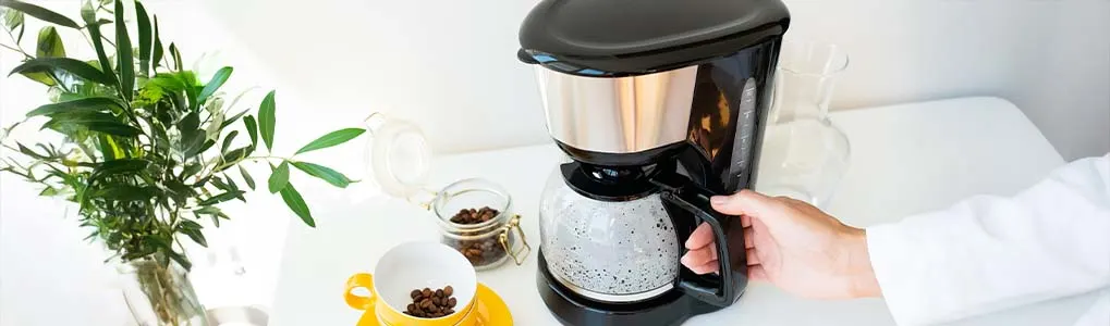 Hand holding the jug of a black filter coffee machine on a white kitchen counter, with a yellow cup, jar of coffee beans, and a small plant nearby.