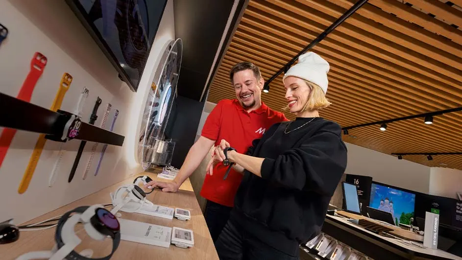 In een winkel staat een vrouw met een muts een horloge te passen, geholpen door een man in een rood shirt.