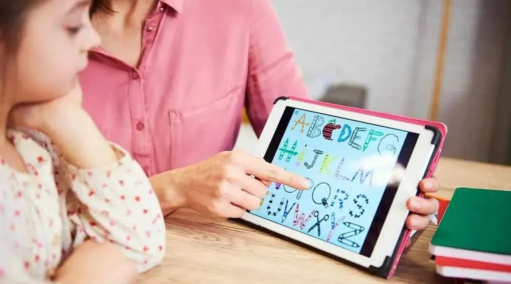 An adult is pointing at a tablet with colourful letters of the alphabet. There are school exercise books on the table. The brand is not visible