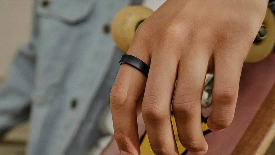 Hand met zwarte ring leunt op een skateboard met gele wielen; een blauw denim kledingstuk op de achtergrond.