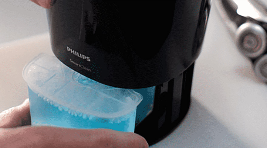 Hand vult blauwe vloeistof in een zwart Philips SmartClean apparaat, met een scheerapparaat op de achtergrond.