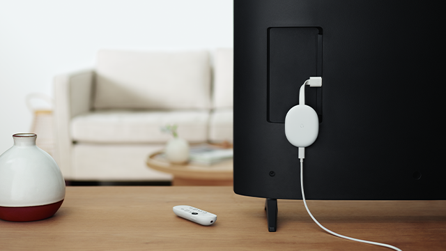 Een zwarte televisie met een witte Chromecast, een witte afstandsbediening en een vaas op een houten tafel.