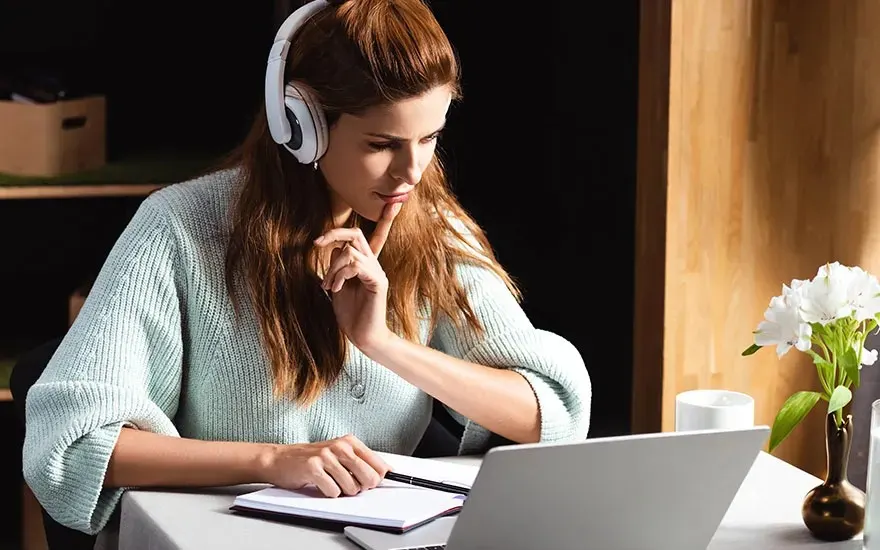 Woman wearing white headphones studies at laptop, taking notes in a notebook at a table with flowers and a coffee mug.