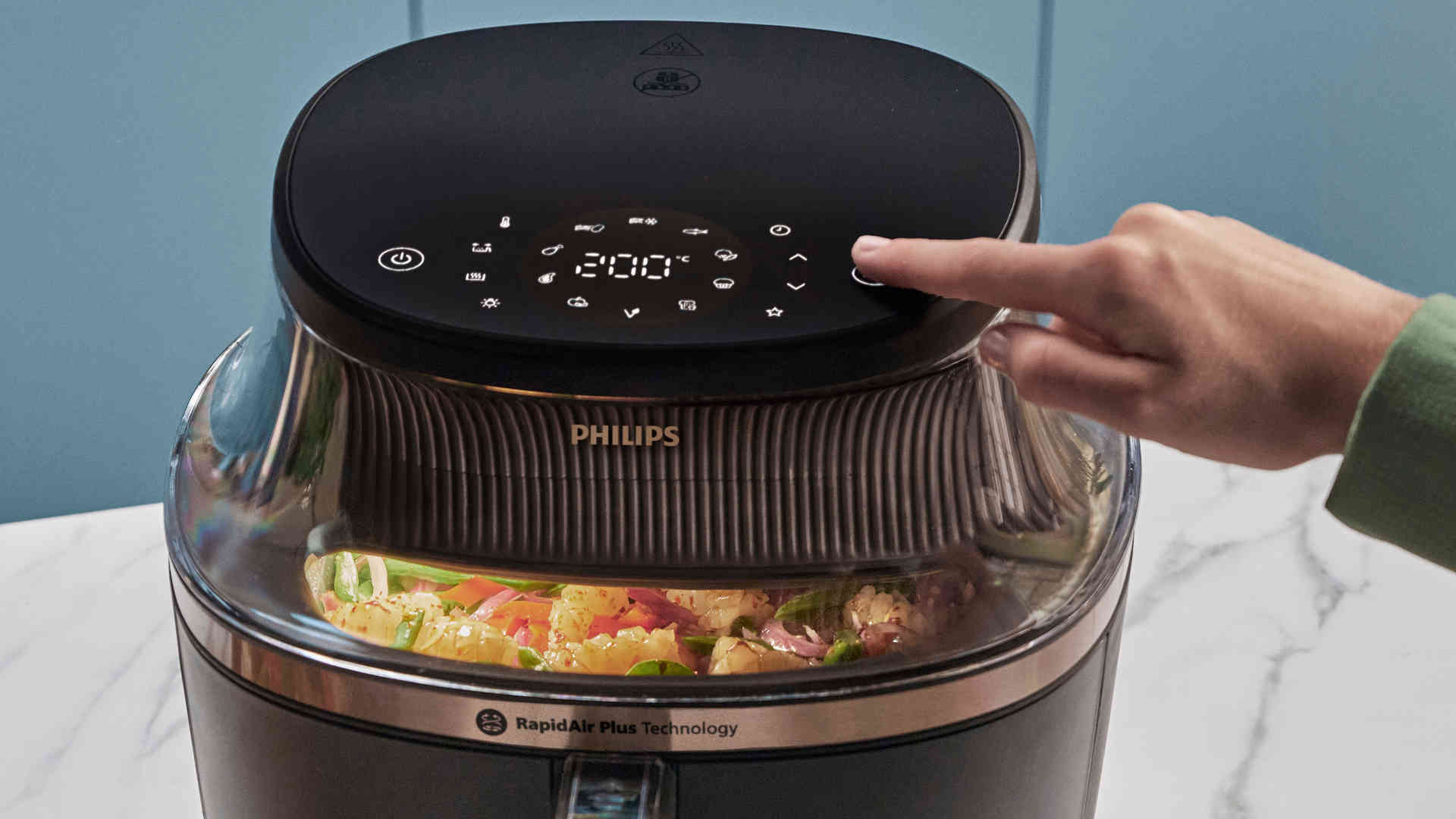 Een Philips airfryer met een hand die een knop bedient, met groenten zichtbaar in het apparaat.