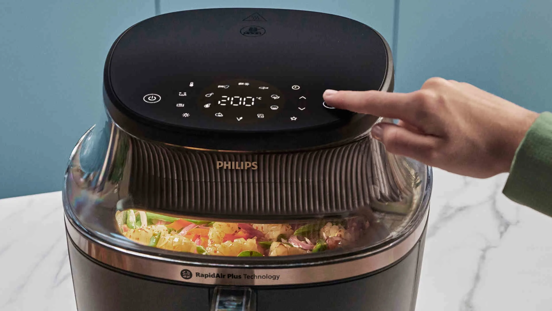 Een Philips airfryer met een hand die een knop bedient, met groenten zichtbaar in het apparaat.
