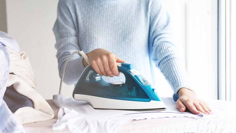 Een persoon strijkt een wit kledingstuk met een blauw en wit strijkijzer, naast een stapel kleding bij een raam.