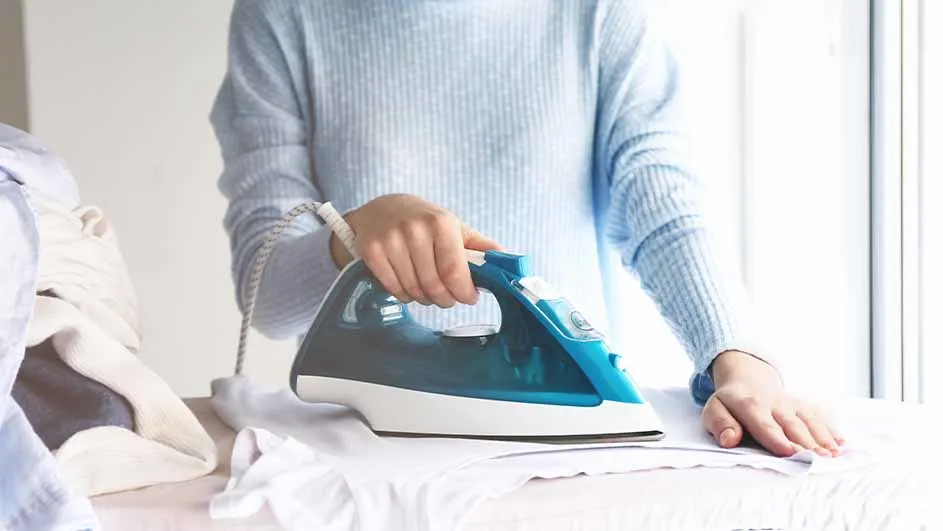 Een persoon strijkt een wit kledingstuk met een blauw en wit strijkijzer, naast een stapel kleding bij een raam.