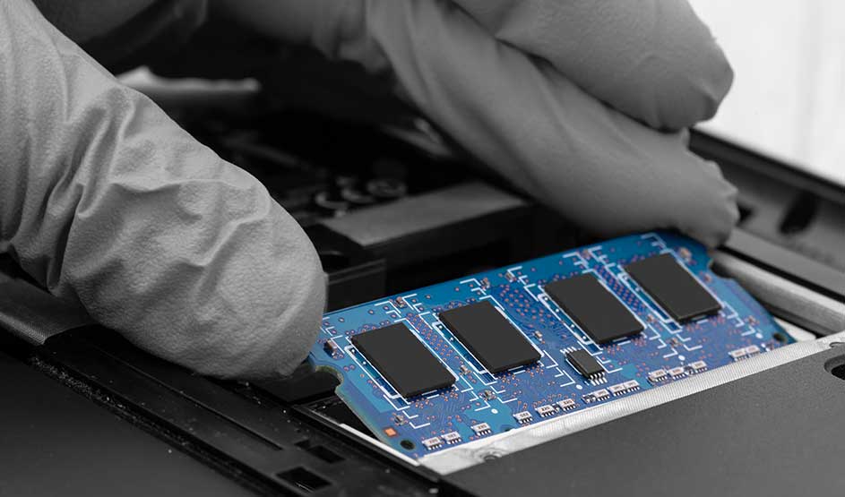 Een hand met een grijze handschoen plaatst een RAM-geheugenmodule in een laptop. Er is geen merknaam zichtbaar op de module.