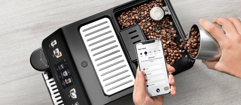 Een hand vult koffiebonen in een zwarte koffiemachine, terwijl een andere hand een smartphone vasthoudt met een app.