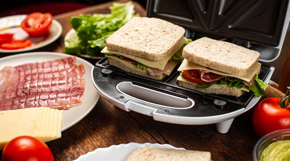 Tosti-ijzer gevuld met ongebakken sandwiches, omringd door verse tomaat, sla, kaas en ham op houten keukenblad, klaar om te grillen.
