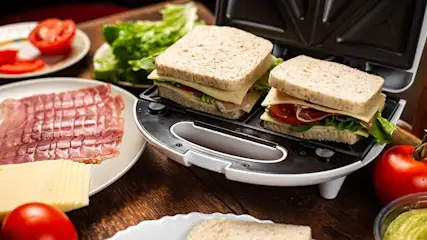 Tosti-ijzer gevuld met ongebakken sandwiches, omringd door verse tomaat, sla, kaas en ham op houten keukenblad, klaar om te grillen.