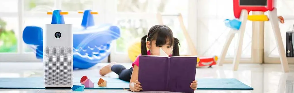 A young girl with pigtails reads a book while lying on a blue mat in a playroom with toys and an air purifier.