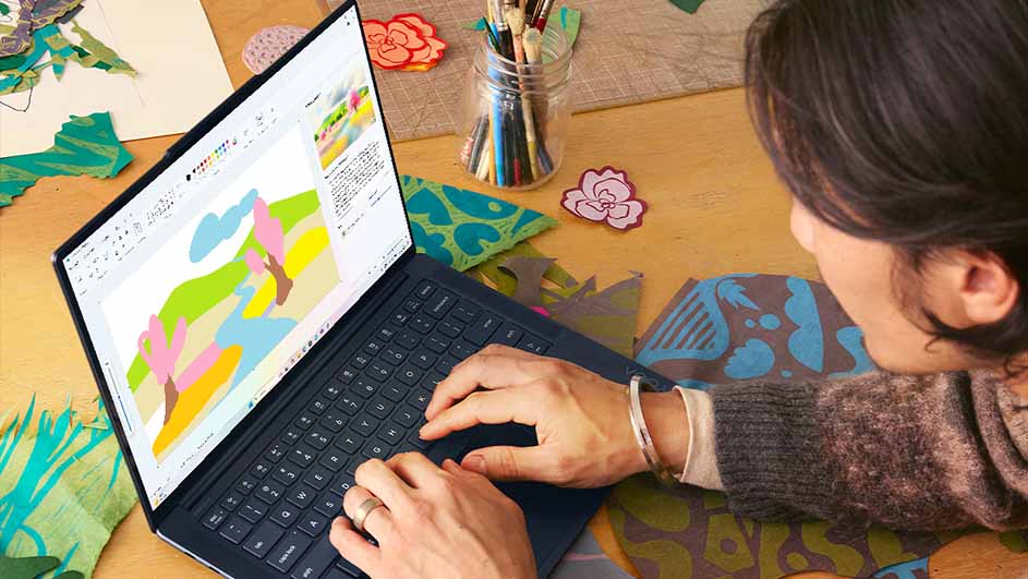 Een vrouw typt op een laptop met een kleurrijke tekening op het scherm, omringd door knutselspullen en decoraties.