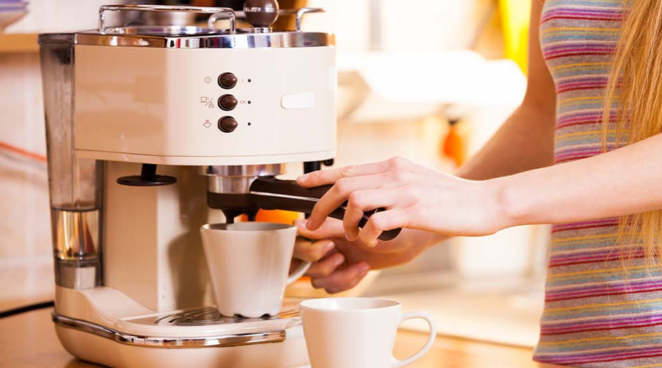 A hand operates an espresso machine with a white cup underneath, ready to brew coffee in a warm kitchen environment.
