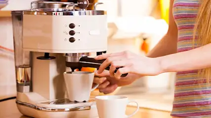 A hand operates an espresso machine with a white cup underneath, ready to brew coffee in a warm kitchen environment.