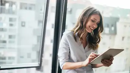 Een vrouw in een blauw hemd gebruikt een tablet bij een groot raam met uitzicht op gebouwen.