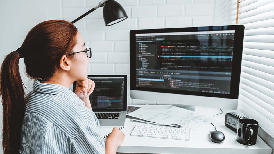Vrouw bekijkt computerschermen met code op een bureau met een toetsenbord, muis, laptop en een mok. Een lamp staat aan de zijkant.