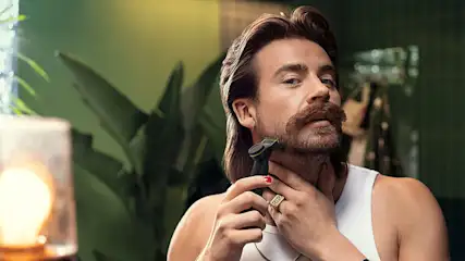 Een man met een snor en een wit hemd gebruikt een zwarte trimmer voor zijn baard, met planten op de achtergrond.