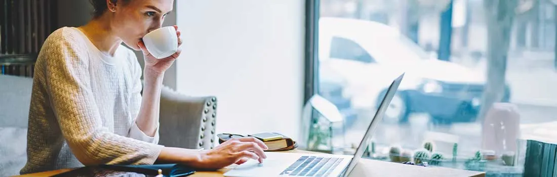 Woman drinking coffee and focused on her laptop near a window in a cozy space.