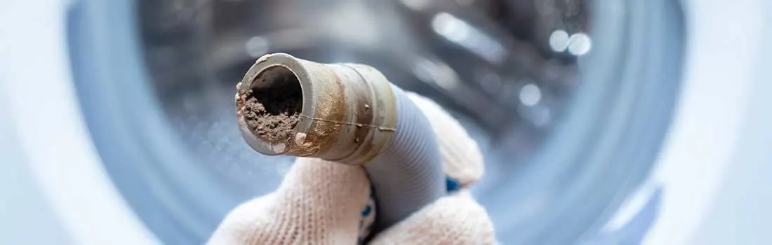 A clogged washing machine drain hose held by a hand wearing a white glove. The pipe is filled with dirt deposits and limescale. In the background, the drum of the washing machine is visible.