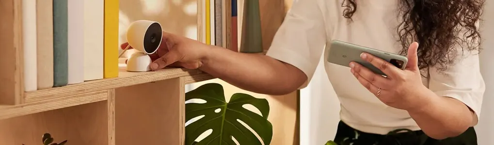 Een vrouw plaatst een witte Google Nest Cam op een plank, terwijl ze haar smartphone vasthoudt. De camera is compact en modern vormgegeven.