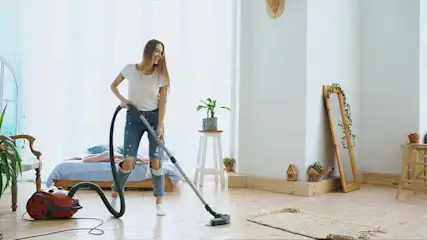 Vrouw stofzuigt de woonkamer met een rode stofzuiger. Er staan planten, een spiegel en een bed in de kamer.