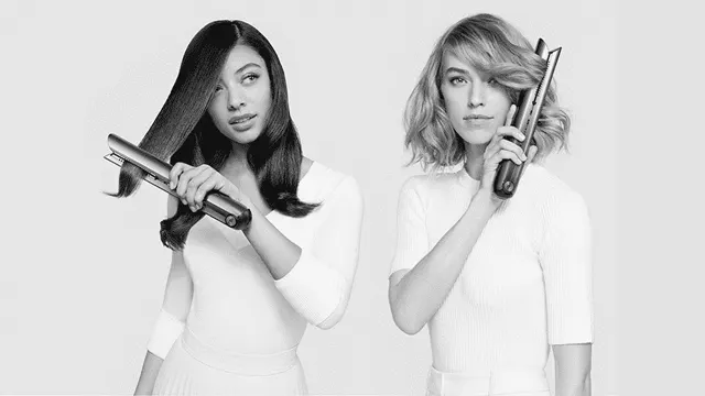 Twee vrouwen in witte shirts. Eén gebruikt een stijltang, de ander houdt er één tegen haar gezicht op een grijze achtergrond.