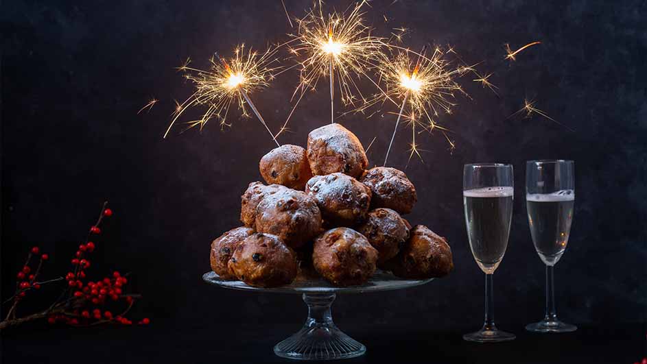 Een stapel oliebollen met poedersuiker en brandende sterretjes op een glazen etagère, naast twee glazen met drank. Rode bessen op de voorgrond.