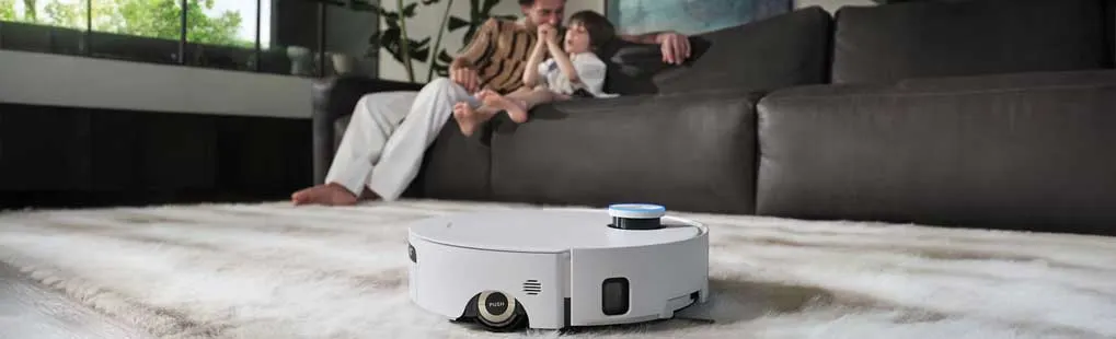 Een robotstofzuiger rijdt over een licht tapijt; op de bank zitten een volwassene en een kind samen te ontspannen in een moderne woonkamer.